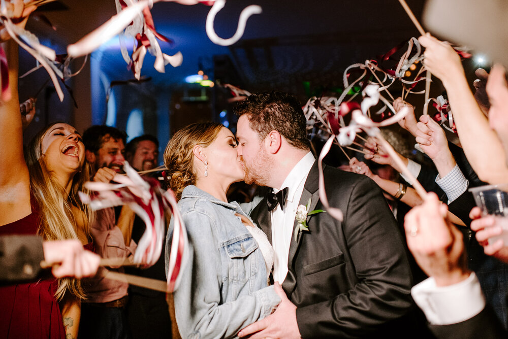 fun wedding exit with streamers and kissing couple with bride wearing reception denim jacket at wilburn street studio by nashville wedding photographer sara bill photography